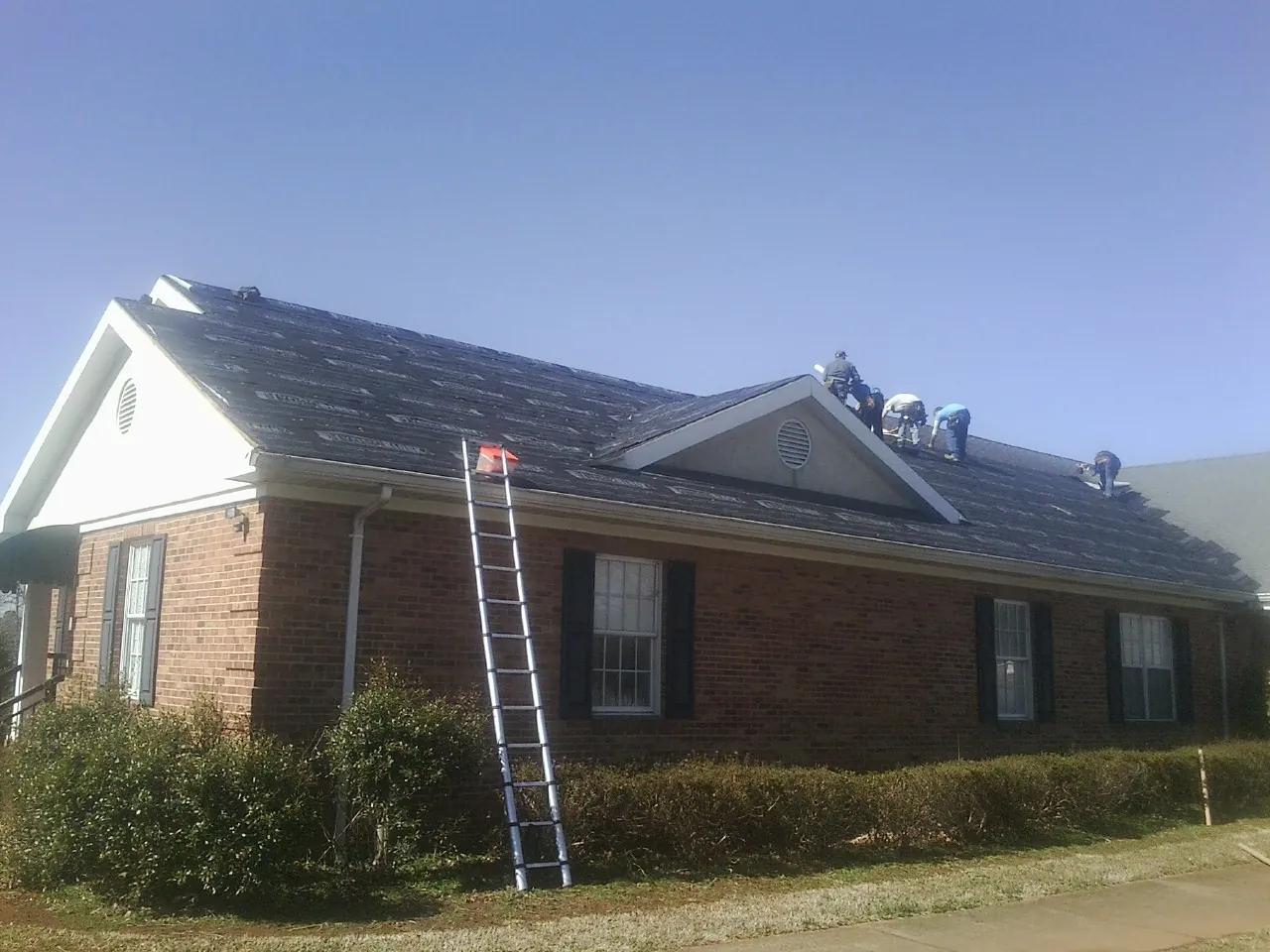 Crew tearing shingles off a brick ranch roof with a ladder set — Metal Roof Repair work by our crew in Goodview